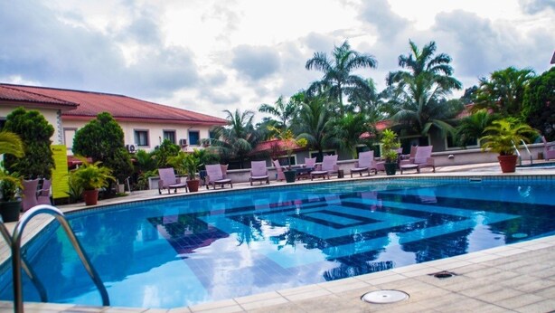 A swimming pool with lounge chairs and potted plants, surrounded by palm trees and a building with a red roof. - Valton Hotels Abuja (Abuja)