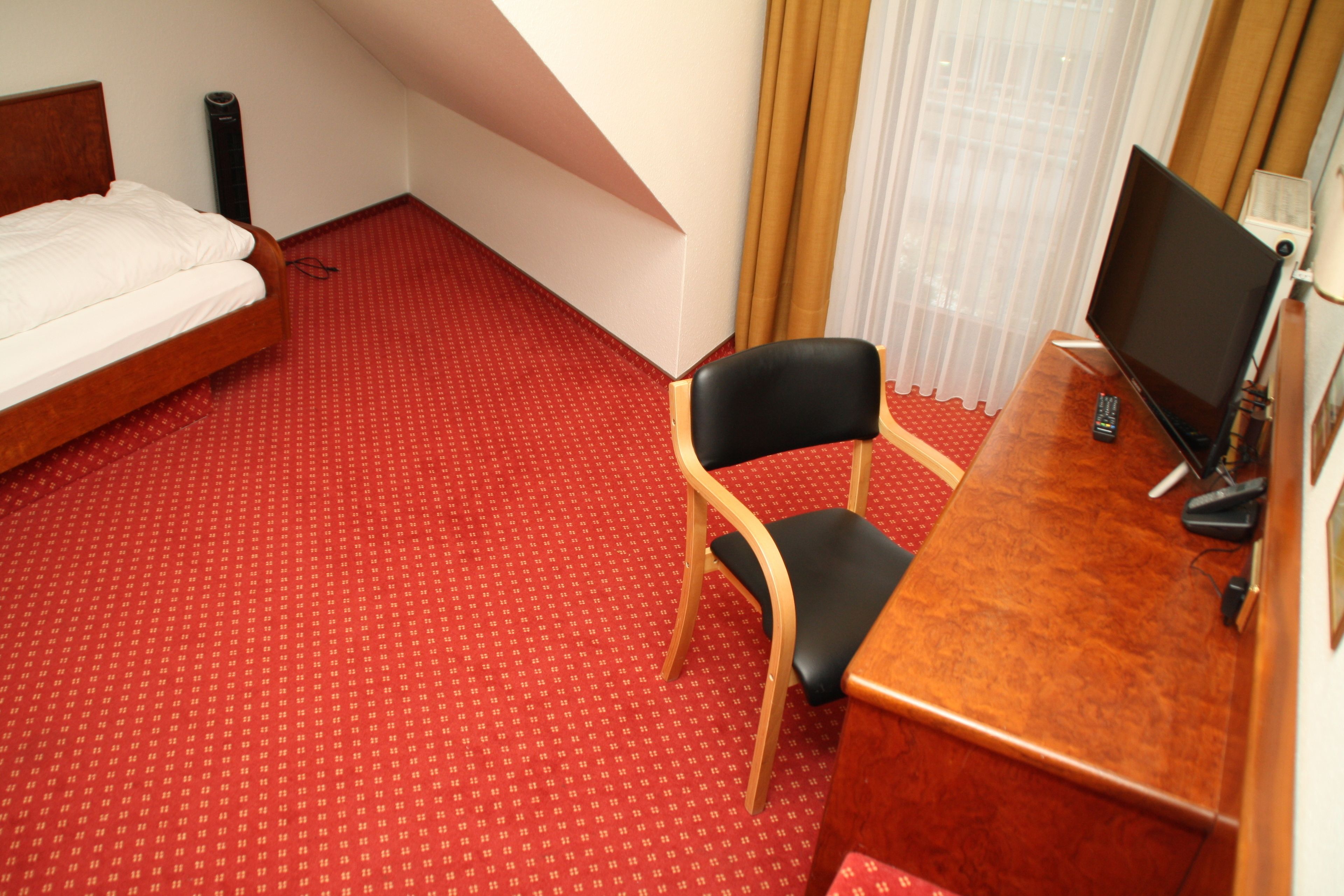 A hotel room with a bed, a desk with a chair, a television, and a red carpeted floor.