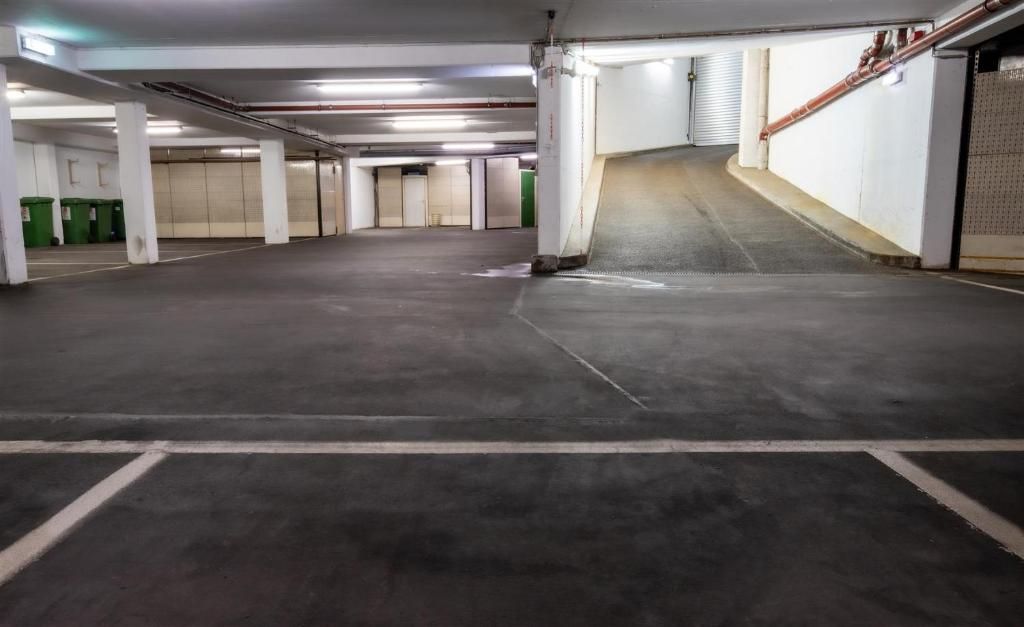 Un estacionamiento vacío con pisos de concreto, pilares blancos y contenedores de basura verdes.