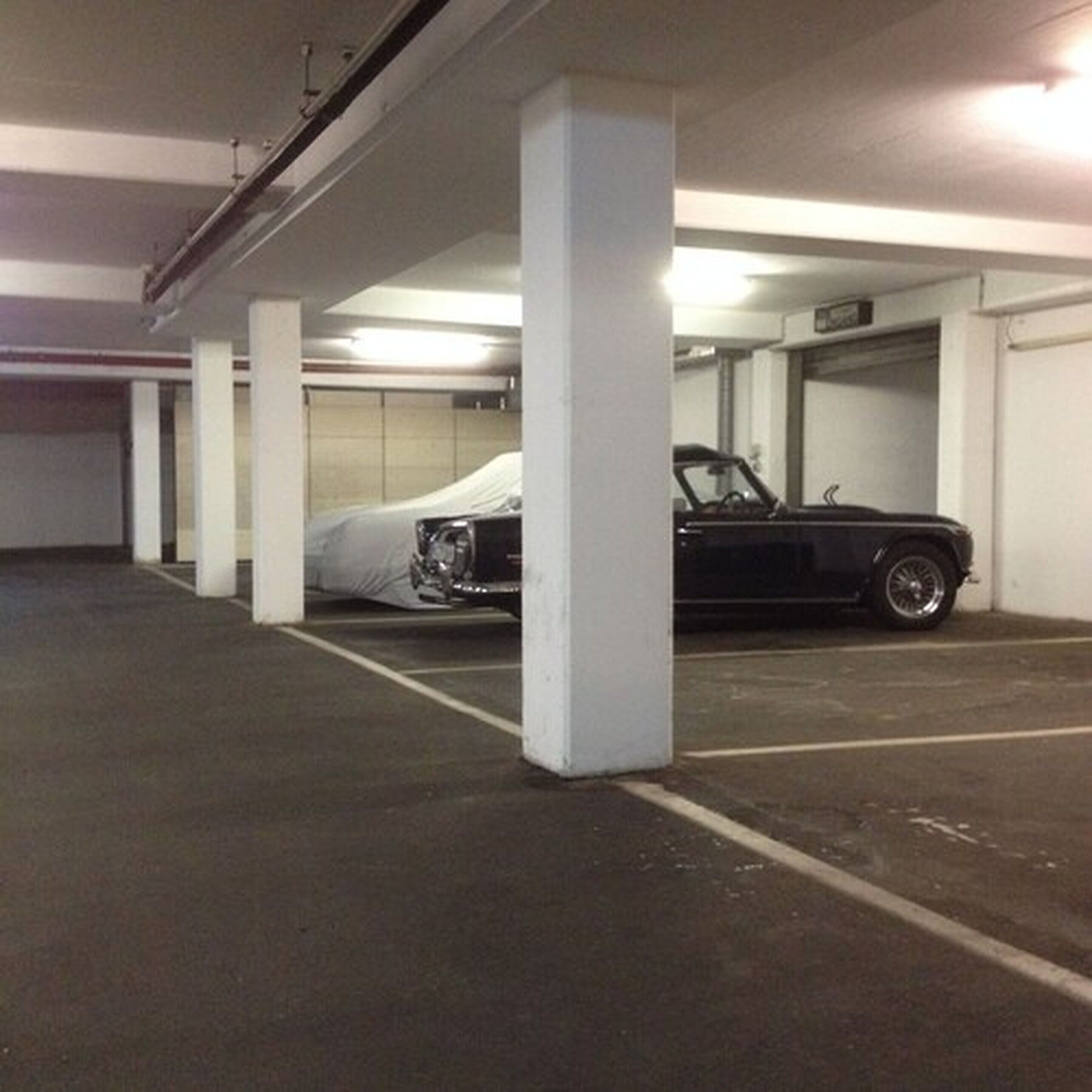 A parking garage with white pillars and a black convertible parked in one of the spaces.
