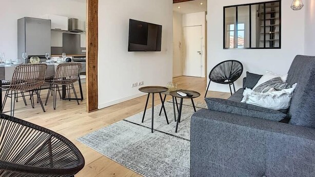 Una sala de estar moderna con un sofá gris, sillas negras, un televisor y una cocina al fondo.