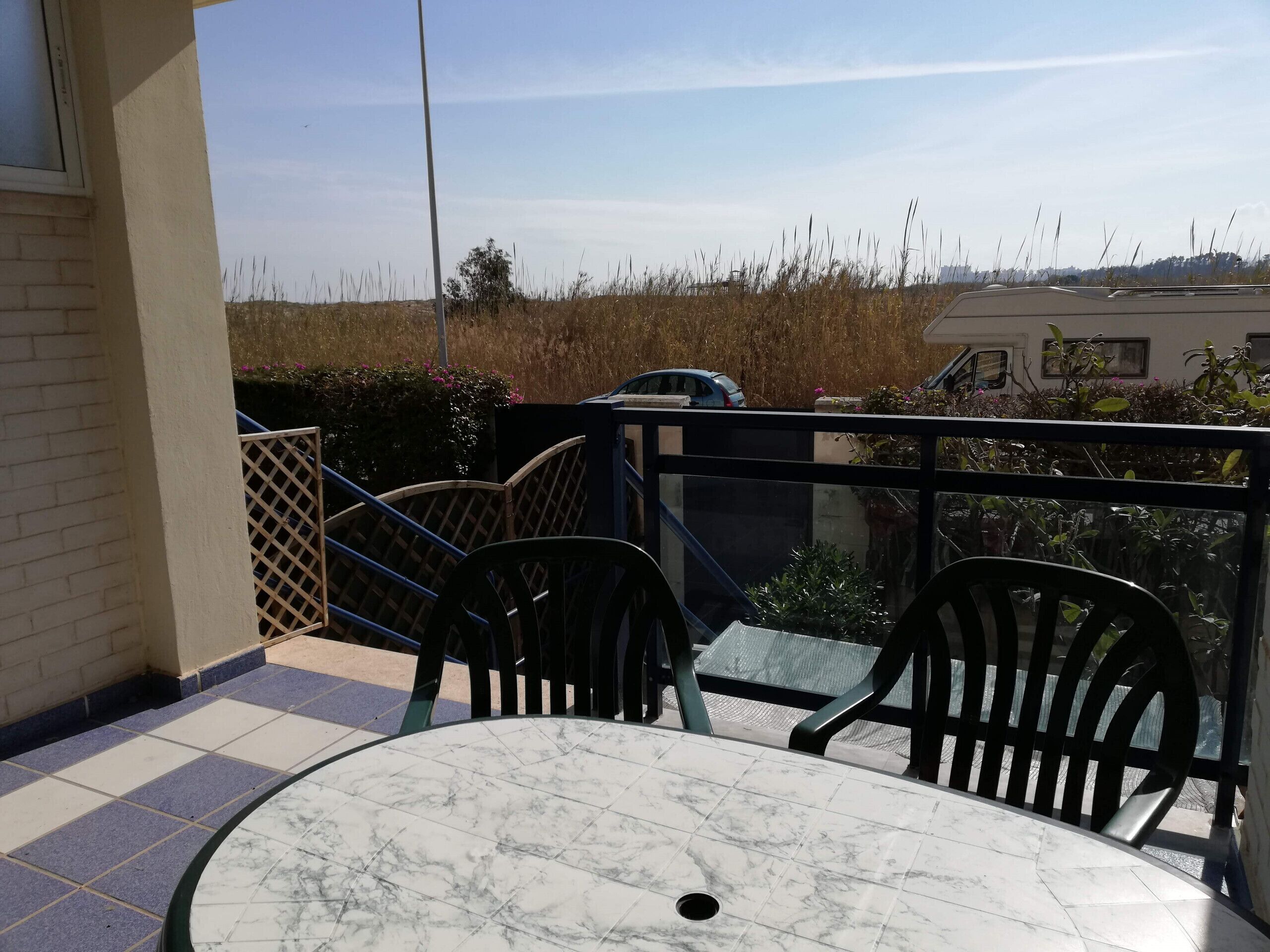 A balcony with a glass table and chairs, a view of a field, and a parked car.