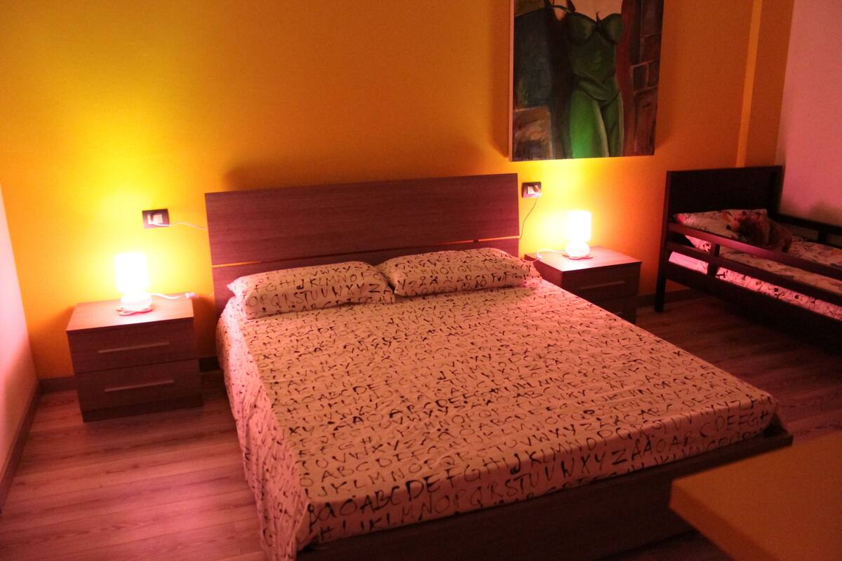 A bedroom with a bed, two bedside tables with lamps, a nightstand, and a bench.