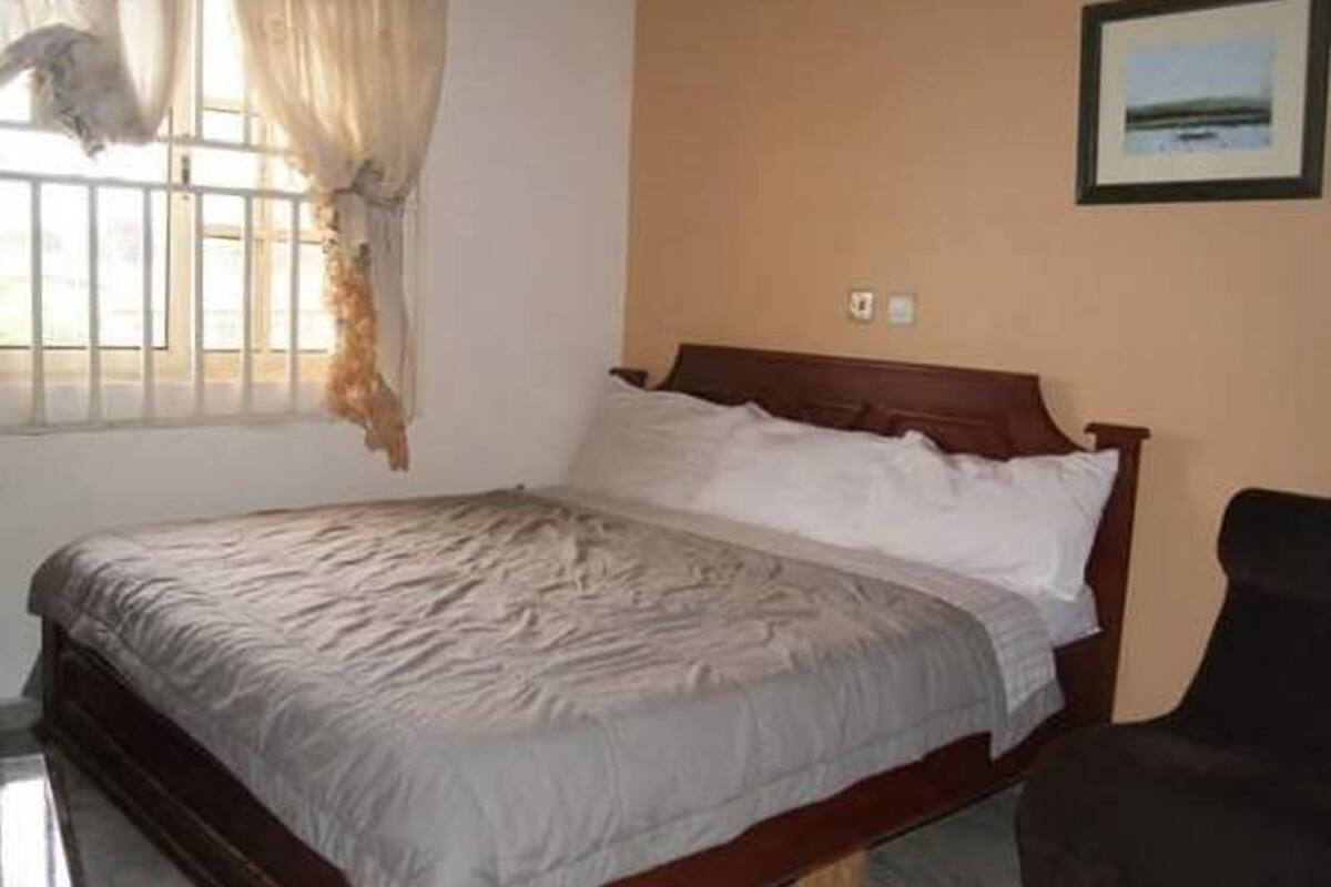 Un dormitorio con cama, ventana con cortinas, interruptor de luz en la pared y un cuadro enmarcado.