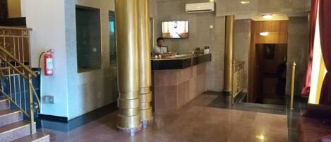 Egy előtér, amelyben található egy recepciós pult, egy tűzoltó készülék, egy lépcsőház és egy nagy oszlop.