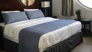 A neatly made bed with a dark headboard, a bedside lamp, a round window with curtains, and a small table with a plant. - Commodore Hotel Abuja (Abuja)