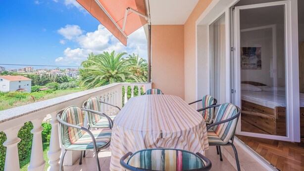 En balkon med et bord og stole, en skydedør der fører til et soveværelse, samt udsigt til palmer og bygninger.