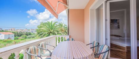 Uma varanda com mesa e cadeiras, uma porta de vidro deslizante que dá para um quarto e vista para palmeiras e edifícios.
