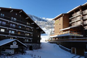 A hotel with multiple balconies, a sign indicating it's 'Hotel Les Arolles,' and a snowy mountain backdrop.