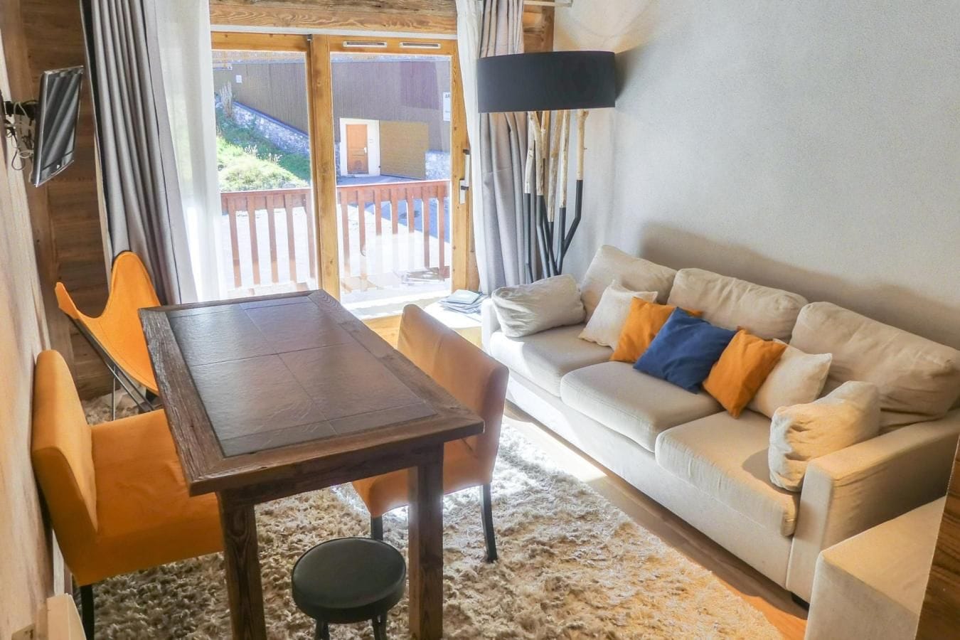 Ein Zimmer mit einem Holztisch, einer Couch mit Kissen, einem Sessel und einem großen Fenster mit Vorhängen.