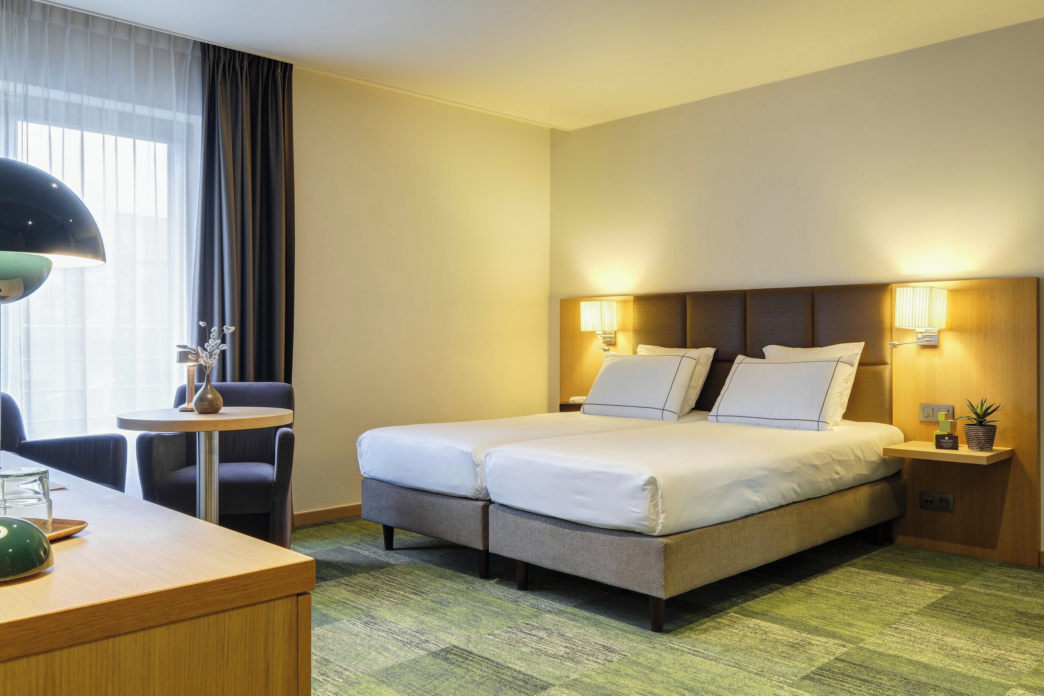 A hotel room with a double bed, two bedside lamps, a small table with a lamp, and a chair.