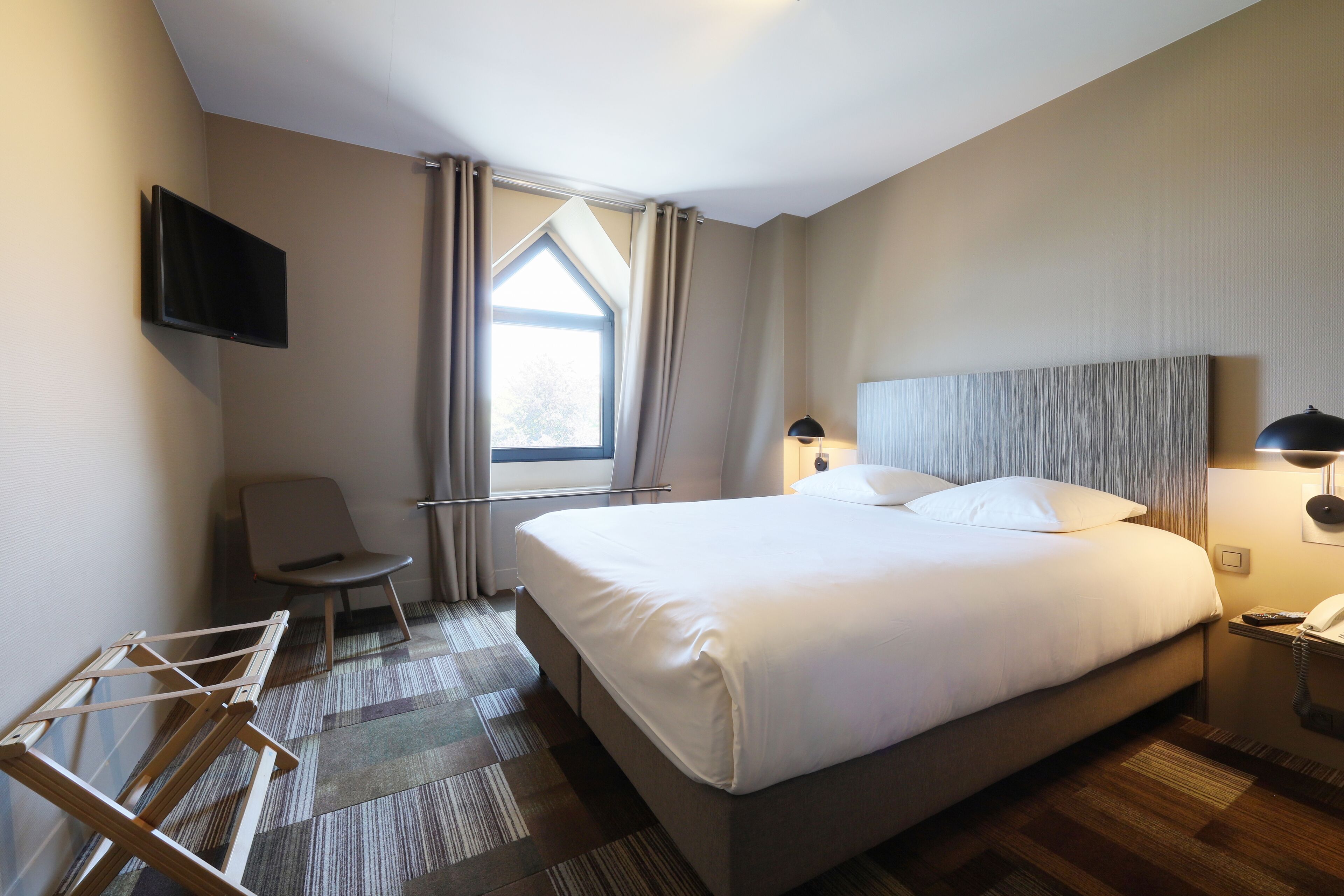 A hotel room with a large bed, a TV mounted on the wall, a chair, a desk with a lamp, and a window with curtains.