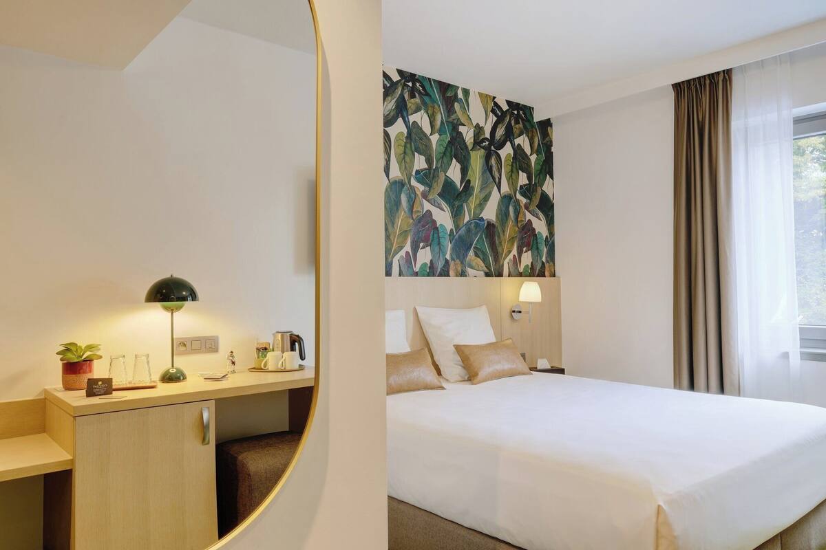 A hotel room with a bed, a desk, a lamp, a coffee maker, a plant, and a large abstract painting on the wall.