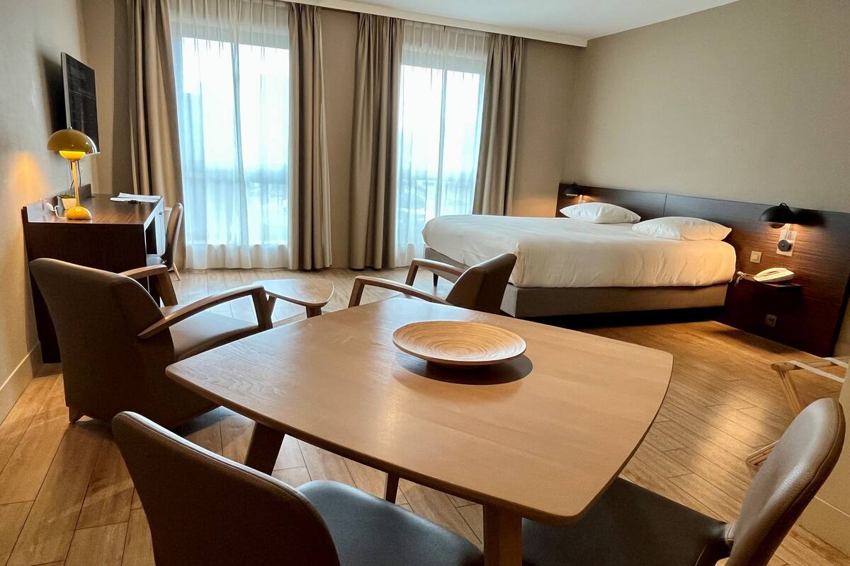 A hotel room with a bed, a desk with a lamp, a dining table with chairs, and a view of the outdoors through large windows.