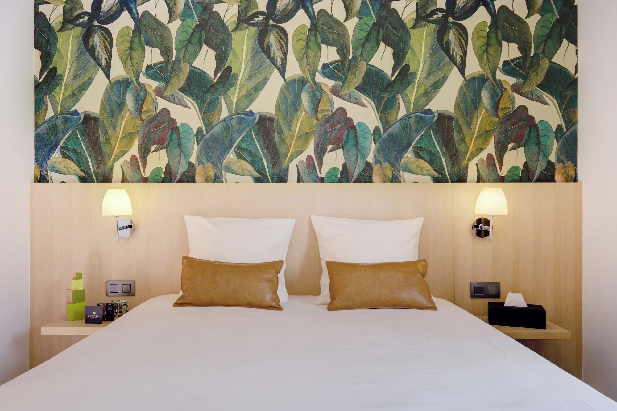 Habitación de hotel con una cama grande, dos almohadas, una mesita de noche con lámpara y una pared con diseño de hojas.
