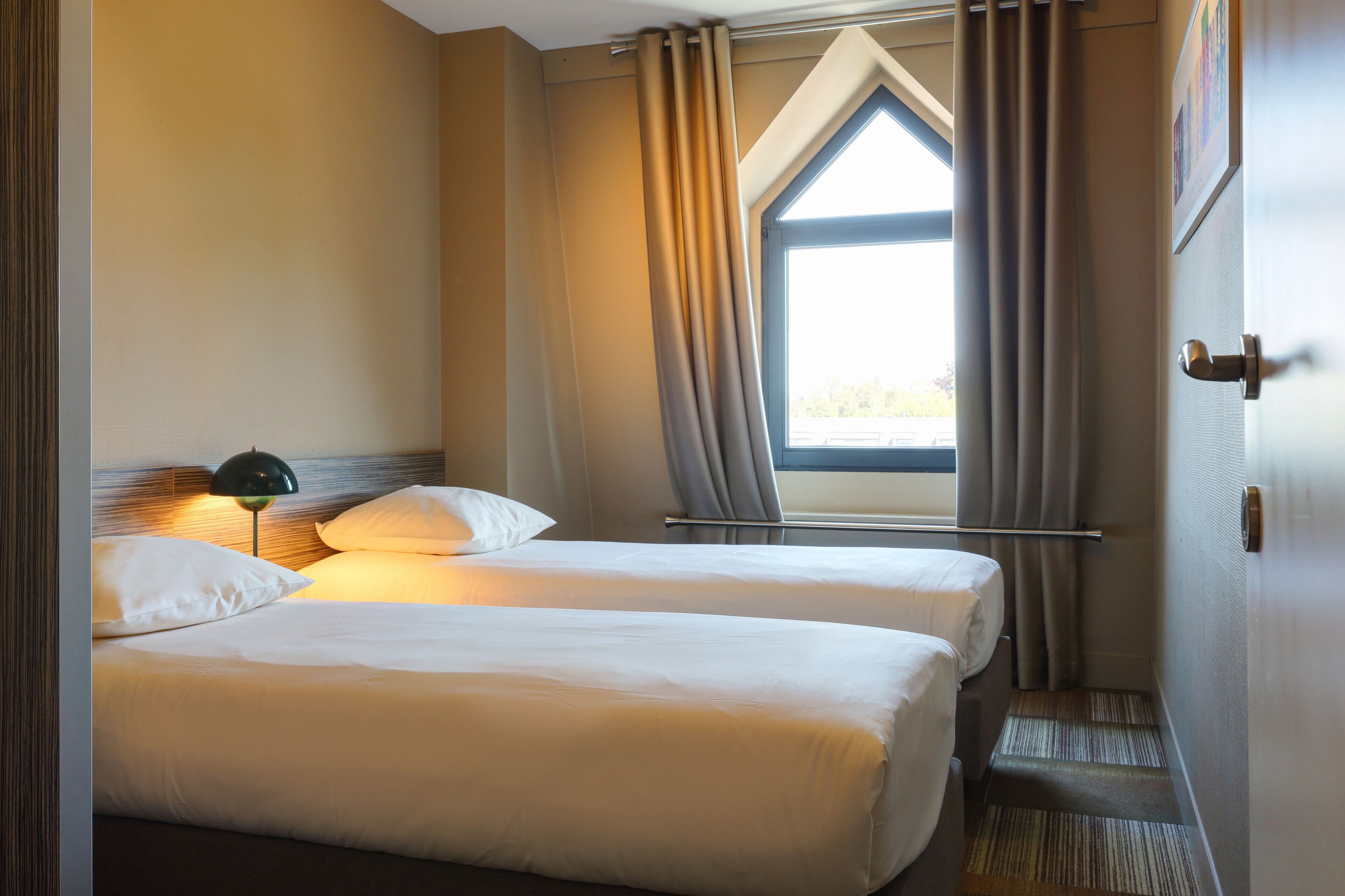 A hotel room with two beds, a window with curtains, a headboard, and a lamp.