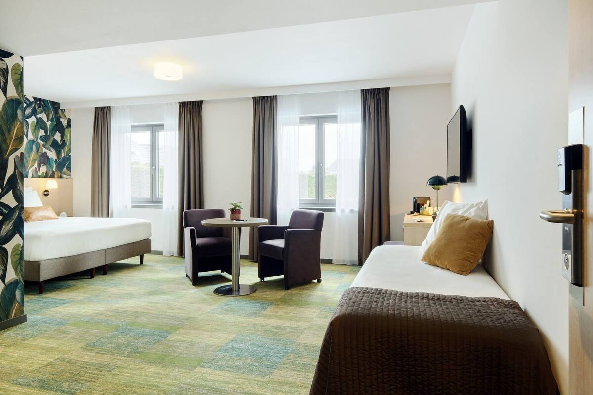 A hotel room with a bed, two armchairs, a small table, a TV, and a window with curtains.