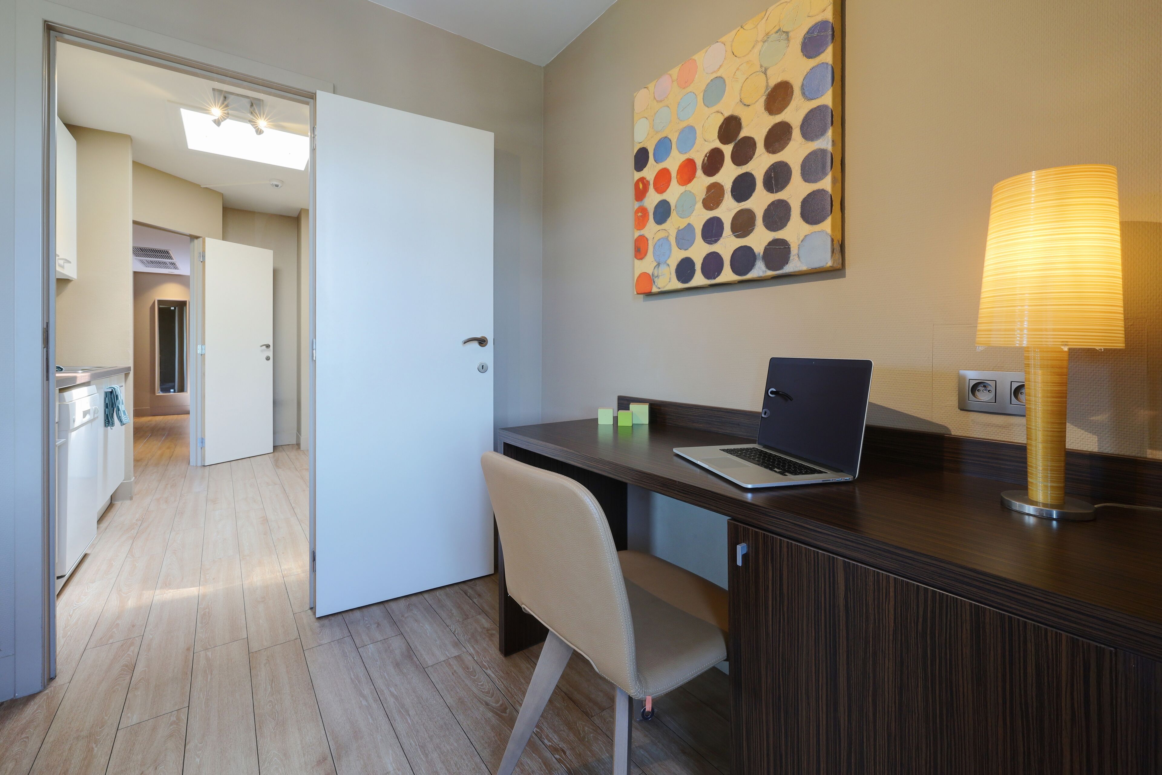 A modern hotel room with a wooden desk, a laptop, a chair, and a wall-mounted lamp.