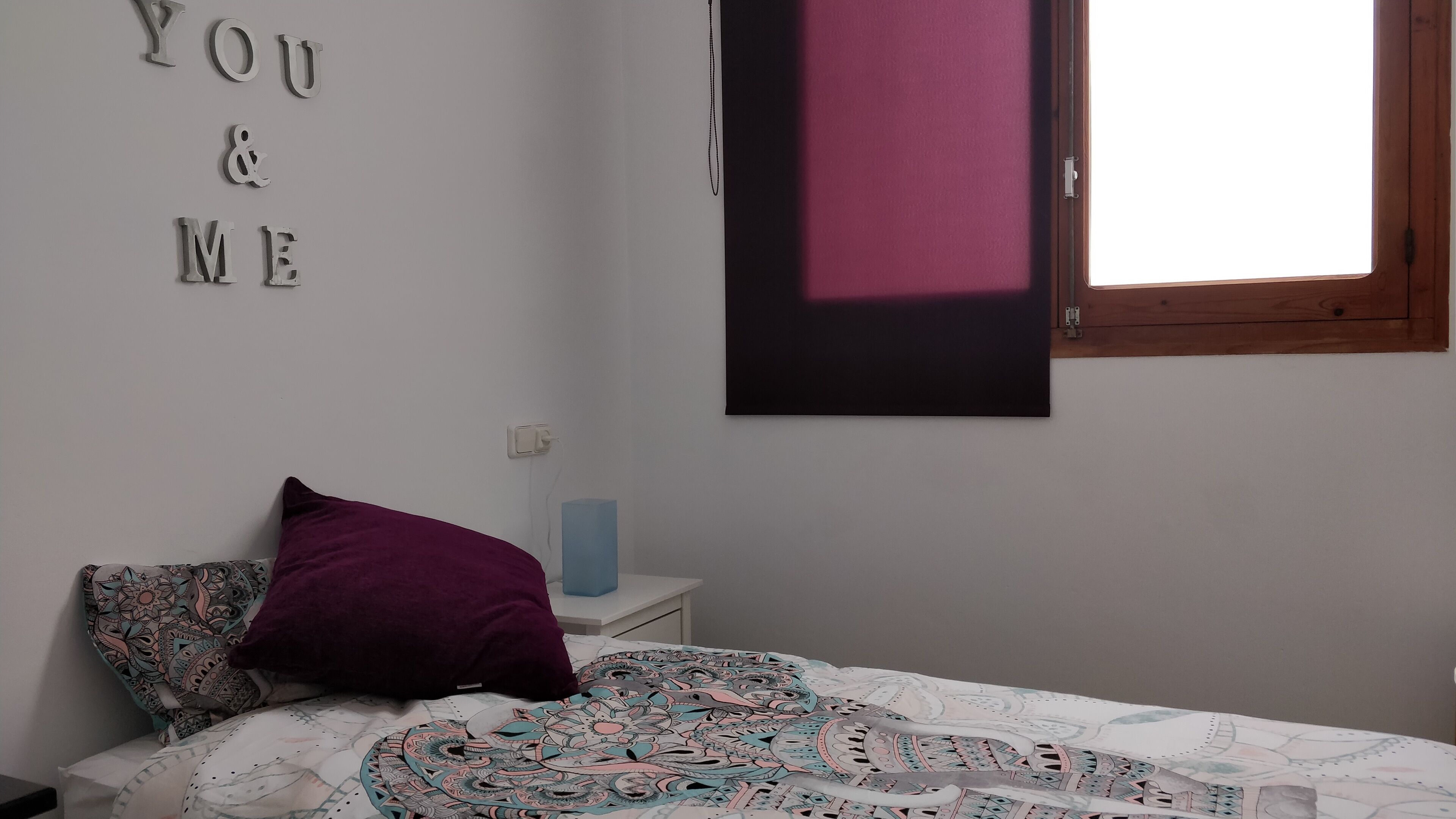 Quarto com cama, almofada roxa, interruptor de luz fixado na parede e janela com moldura de madeira.