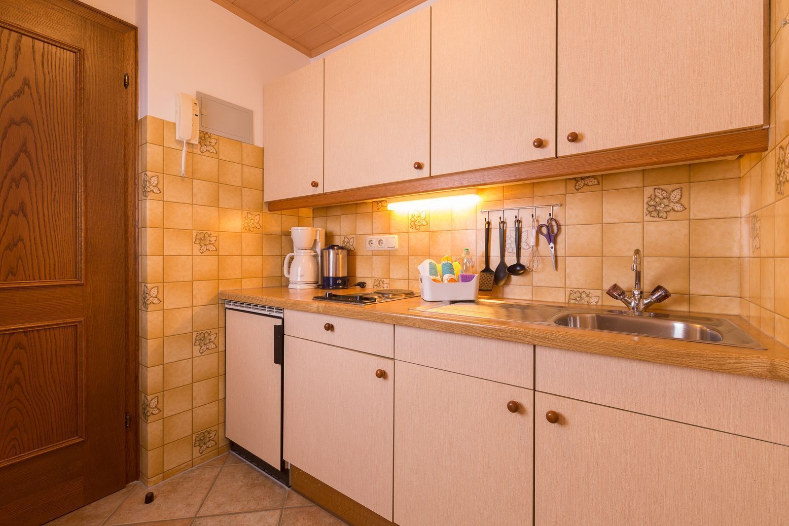 A compact kitchen with wooden cabinets, a built-in dishwasher, and a countertop with a coffee maker, kettle, and utensils.