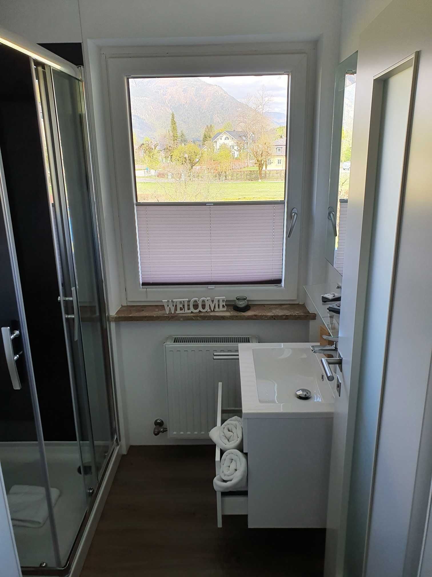 Een moderne badkamer met een glazen douchecabine, een raam met uitzicht op een groen landschap en een wit wastafelmeubel met een handdoekhouder.