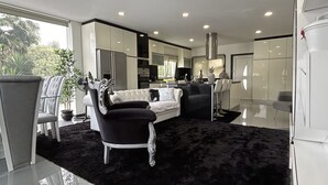 Una sala de estar moderna con un alfombra mullida negra, un sofá blanco y un sillón negro.  Hay una zona de cocina con armarios blancos y un refrigerador de acero inoxidable.