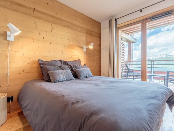Un dormitorio moderno con cabecera de madera, una cama grande con ropa de cama gris, dos lámparas de noche y un balcón con mobiliario exterior.