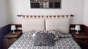 Een hotelkamer met een bed, voorzien van een beddengoed met een patroon, twee kussens met een geometrisch ontwerp, een hoofdbord met een horizontale stang, en een ingelijste foto van een stadszicht aan de muur.