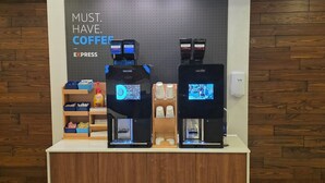 A coffee station with two espresso machines, a display of disposable cups, and a sign that reads "MUST. HAVE. COFFEE."