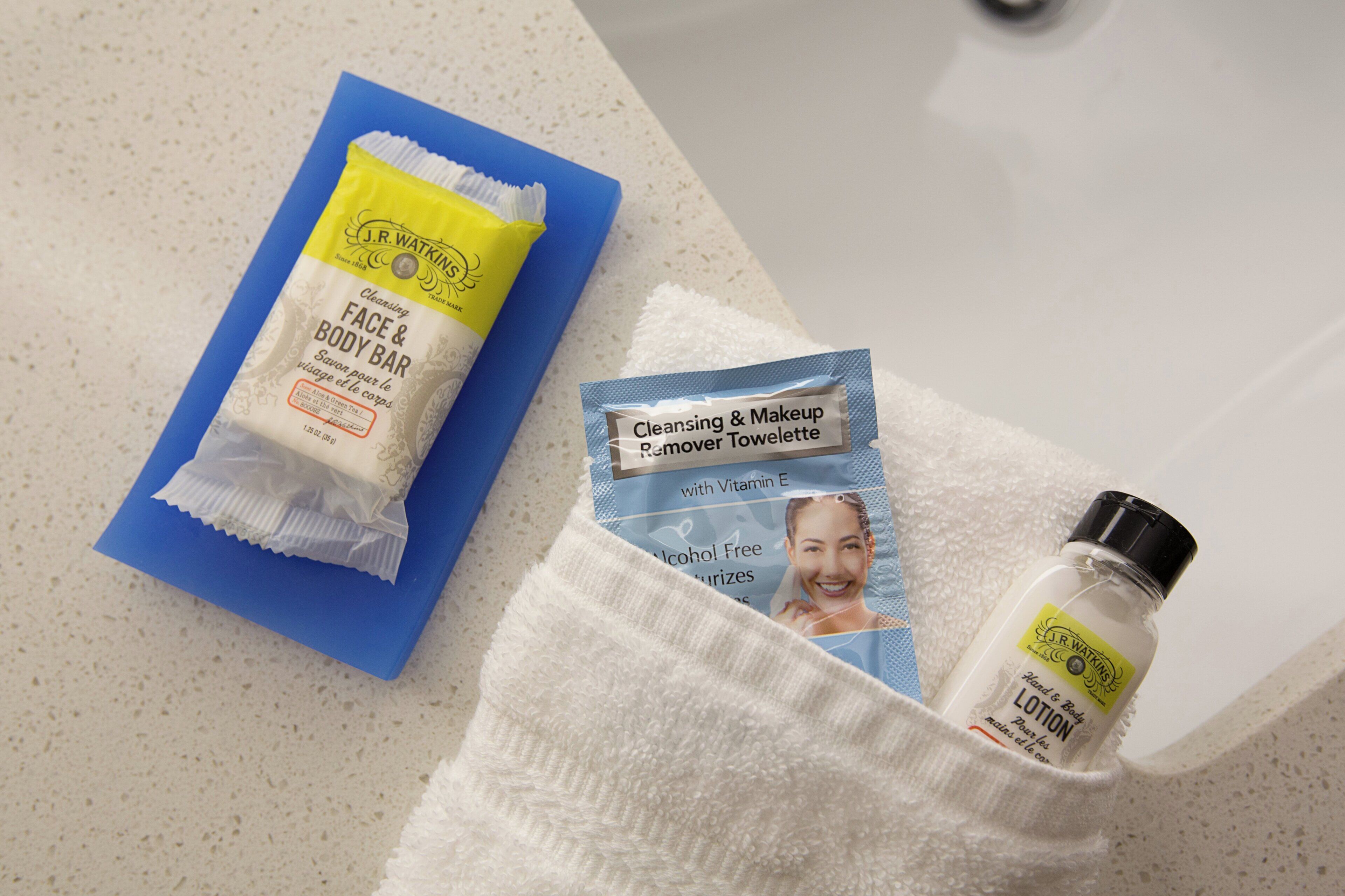 Un baño con un lavamanos blanco, una toalla azul, una pastilla limpiadora J.R. Watkins, un folleto azul y una botella de loción J.R. Watkins.