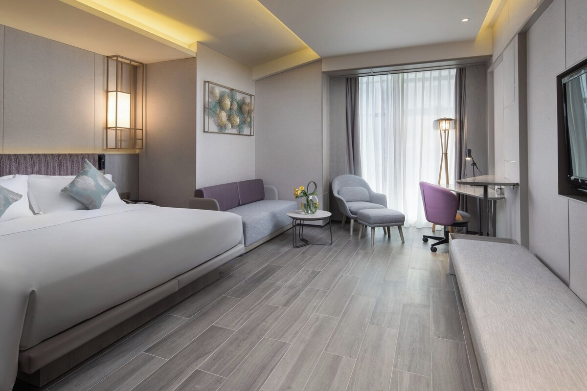 A modern hotel room with a large bed, a sofa, a small table with a vase, a chair, and a desk.