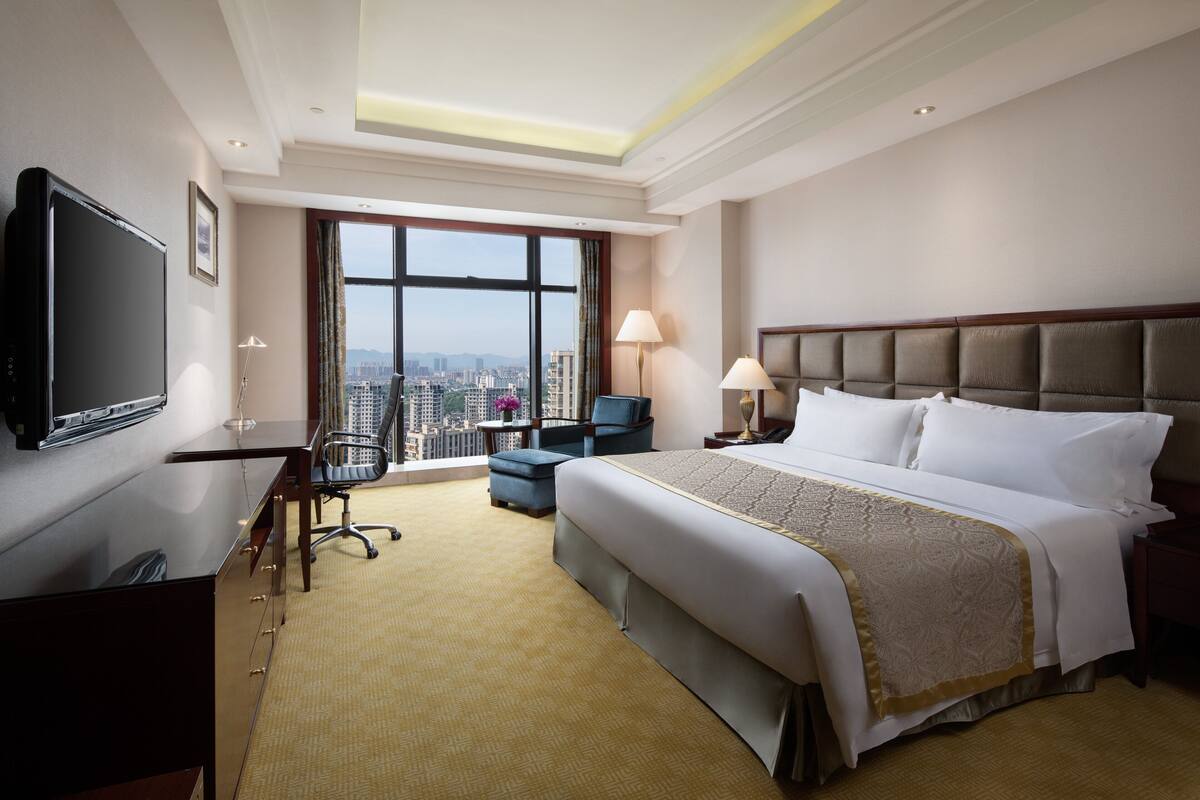 Habitación de hotel con una cama grande, un escritorio con televisor, una silla y vistas a la ciudad a través de amplios ventanales.