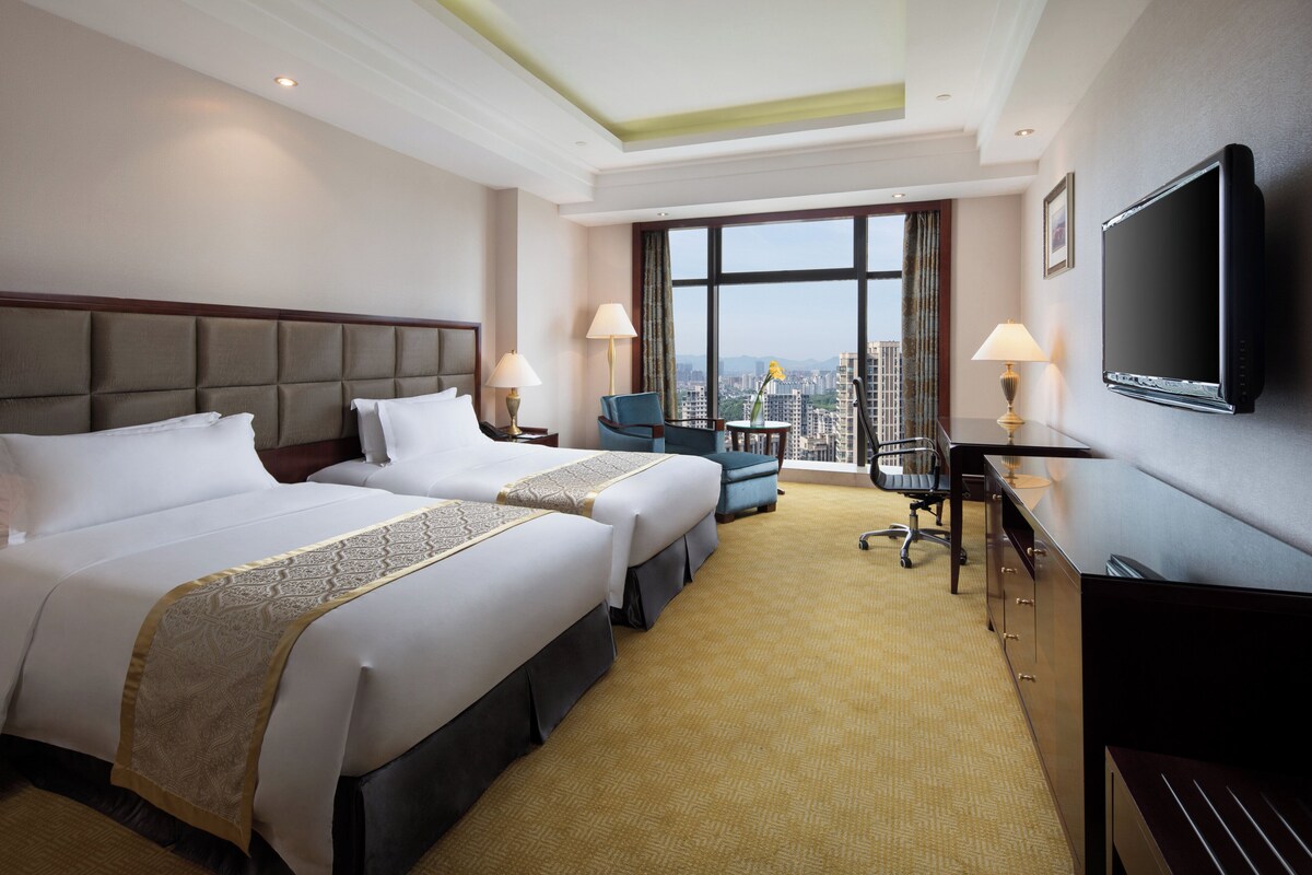 A hotel room with two beds, a desk, a chair, a TV, and a large window offering a city view.