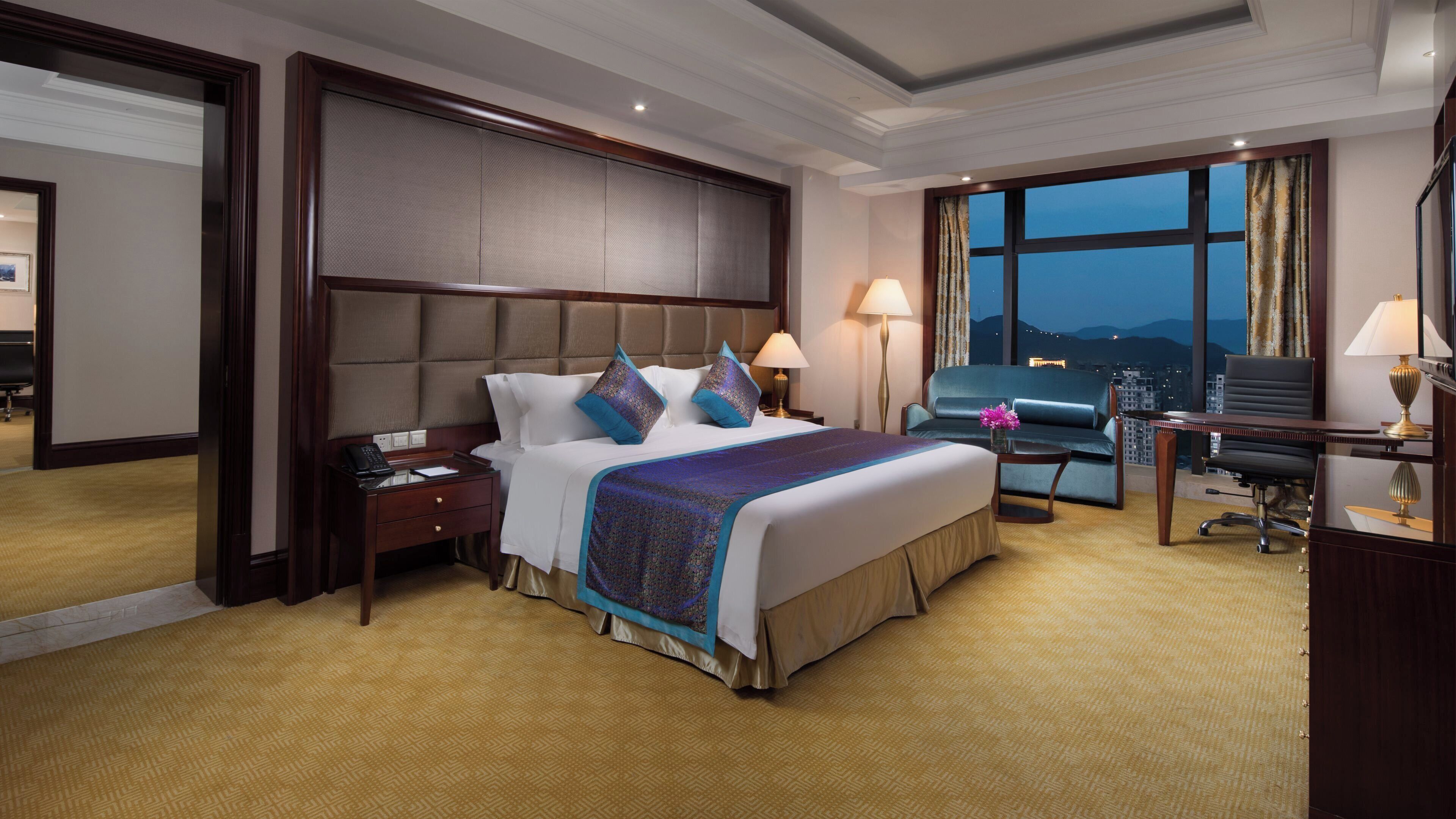 Habitación de hotel con una cama grande, mesitas de noche, un escritorio con una silla y vistas a la ciudad al atardecer.