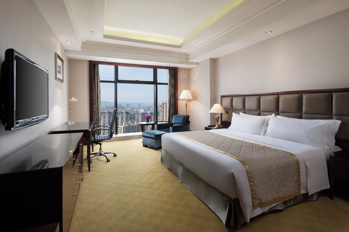 A hotel room with a large bed, a desk, a television, and a view of the cityscape.
