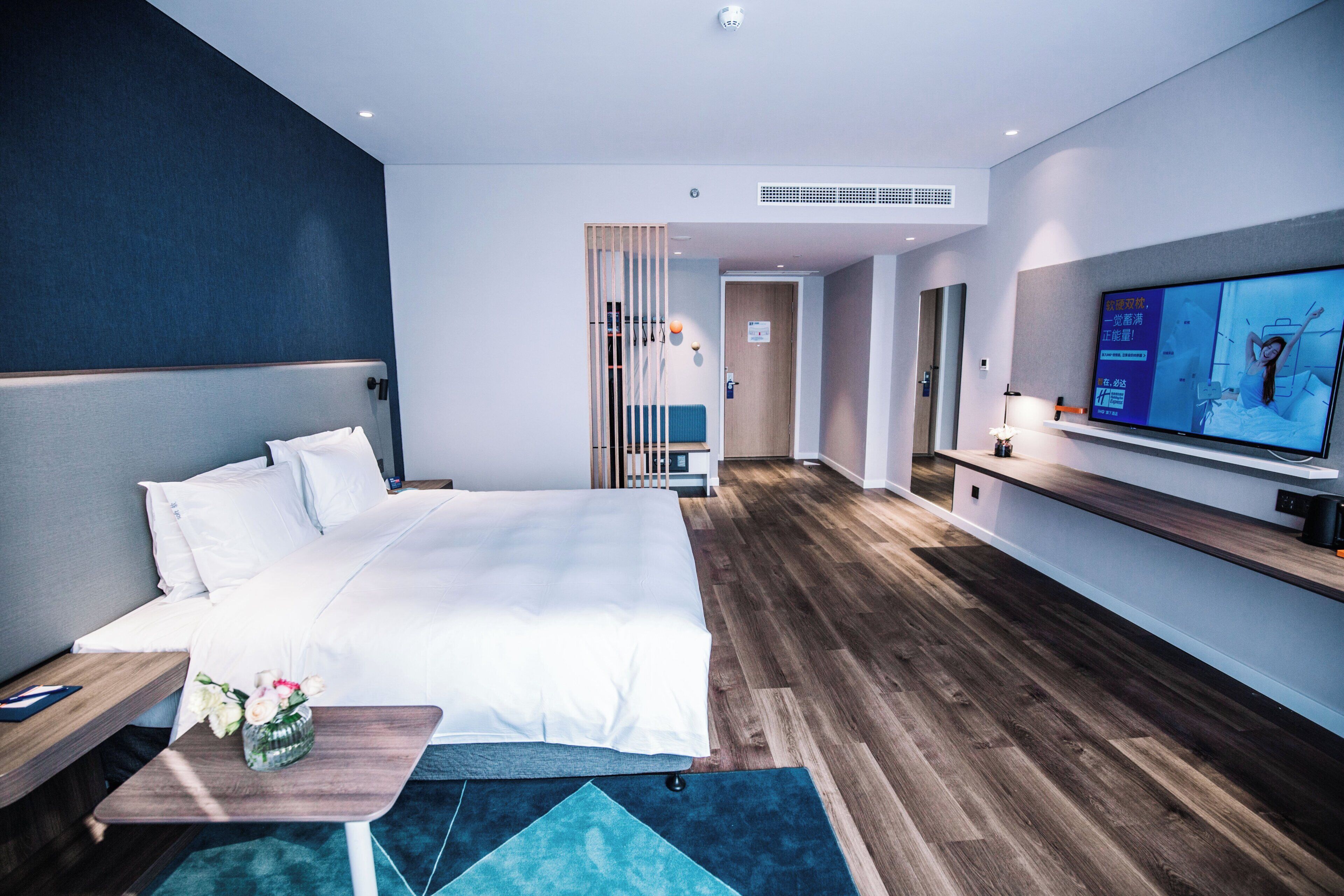A modern hotel room with a large bed, a TV, a desk, and a bathroom visible in the background.