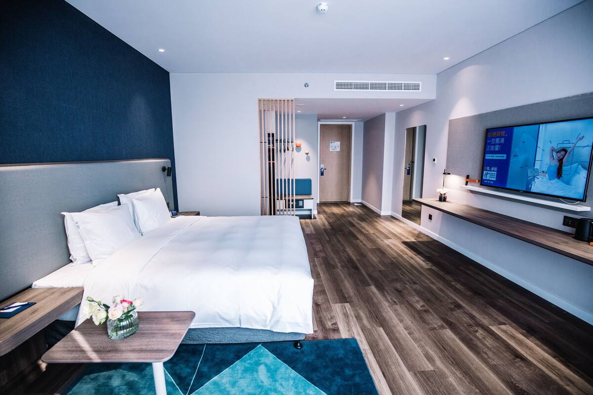 A modern hotel room with a large bed, a TV, wooden flooring, and a small table with a vase of flowers.