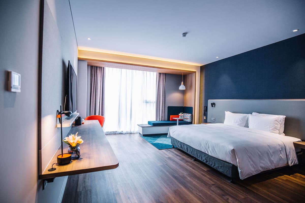 A modern hotel room with a large bed, a desk with a lamp, a TV, and a view of the outdoors.