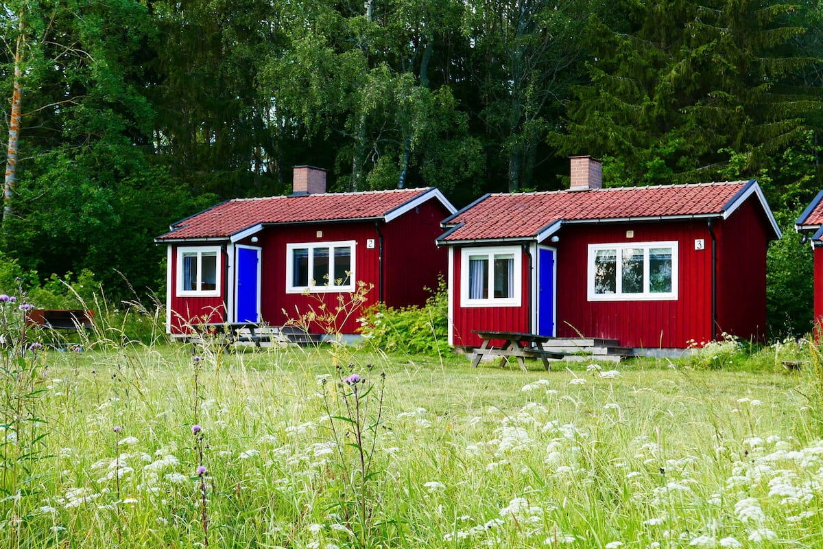 Two red cabins with white trim, numbered 2 and 3, set in a grassy field with wildflowers and trees in the background.