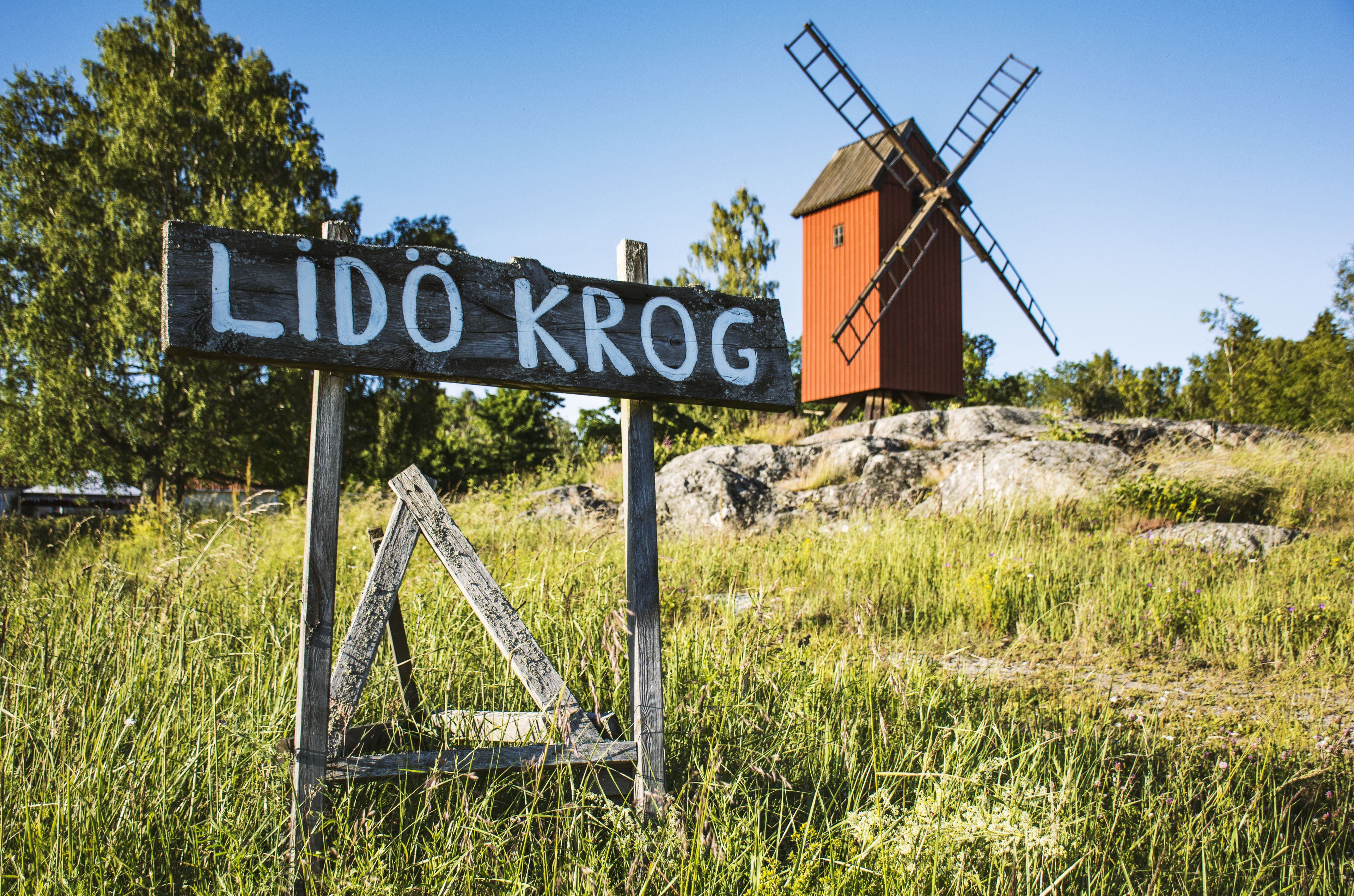 Une enseigne en bois portant l’inscription « LIDÖ KROG » et, en arrière-plan, un moulin à vent.