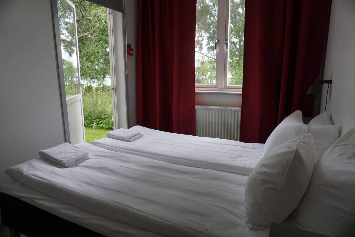 A room with two beds, red curtains, a window with a view of greenery, and a white radiator.
