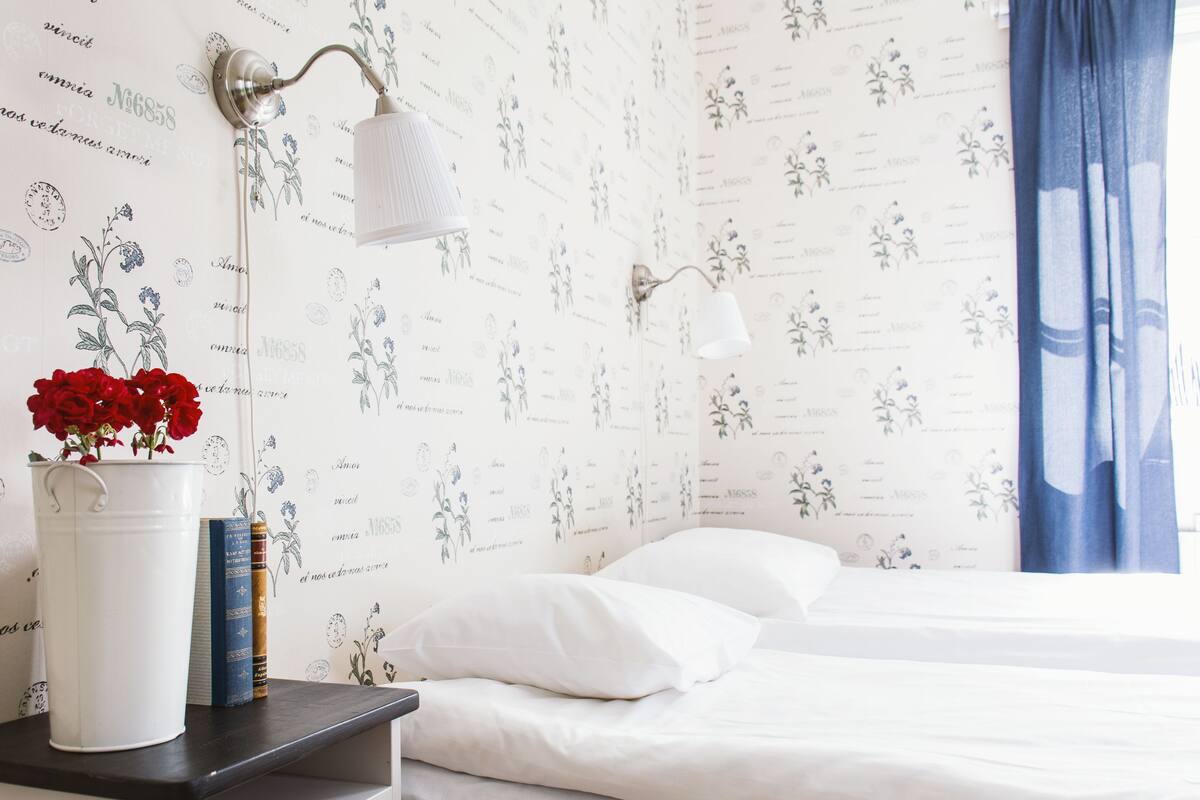 A bedroom with a bed, white bedding, a nightstand with a vase of red flowers, and a wall with floral wallpaper.