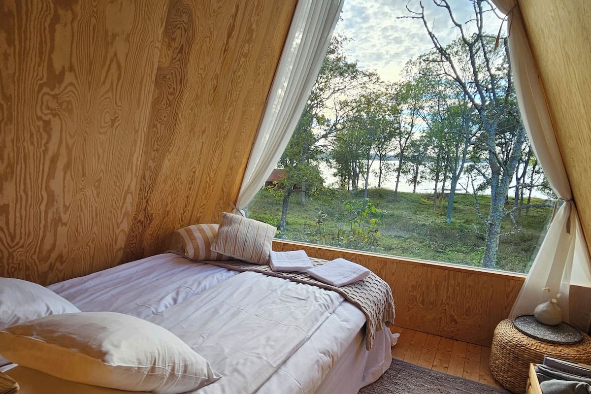 A cozy bedroom with a large window, wooden walls, a bed with white linens, and a view of trees and a lake.