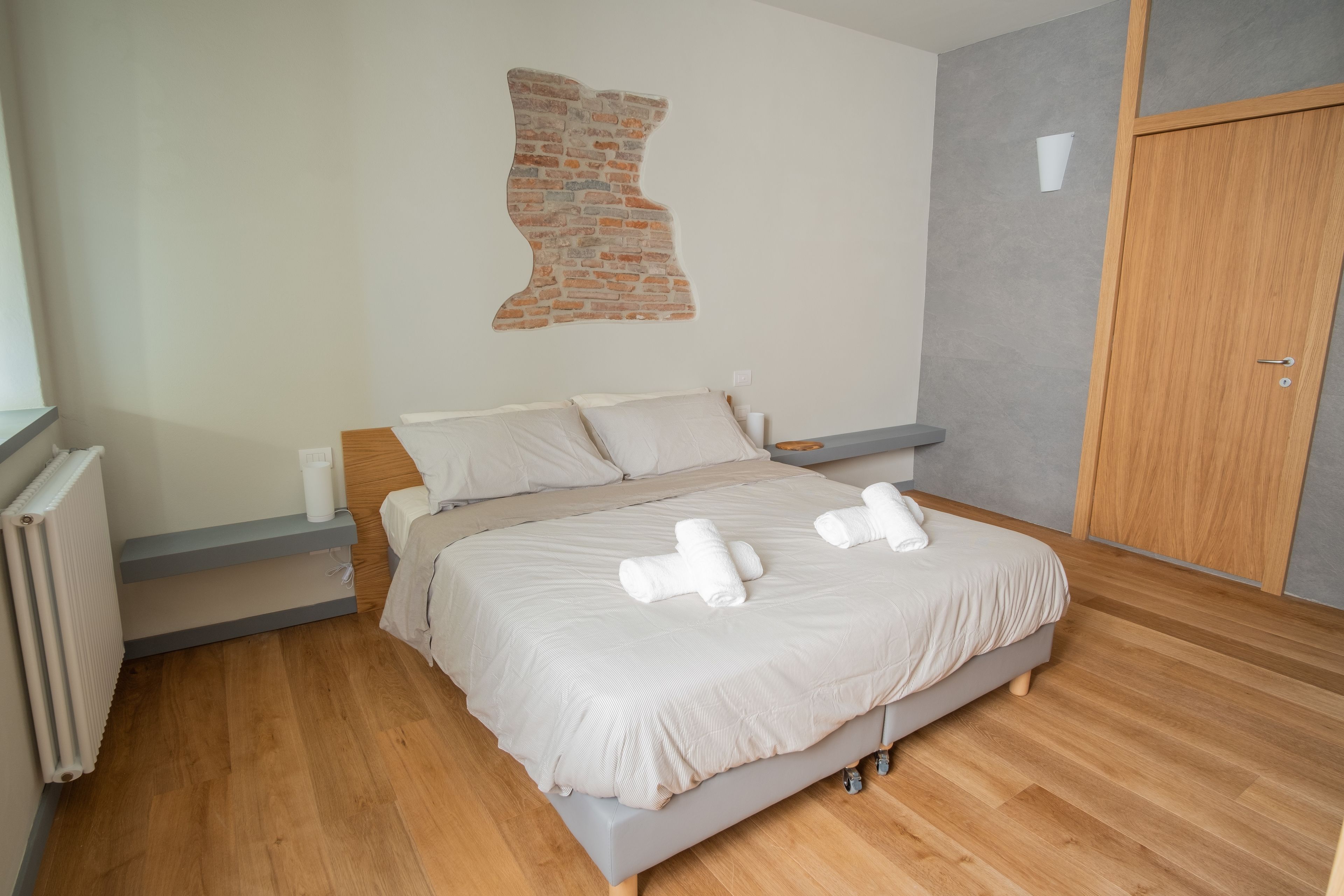 A modern bedroom with a bed, wooden floor, a brick accent wall, and a wooden wardrobe.