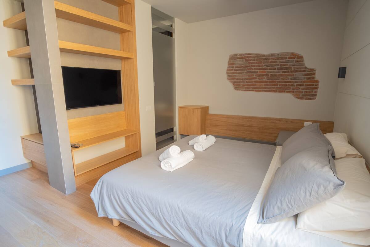 A modern bedroom with a bed, a TV mounted on a wooden shelf, a brick wall accent, and neatly folded towels.