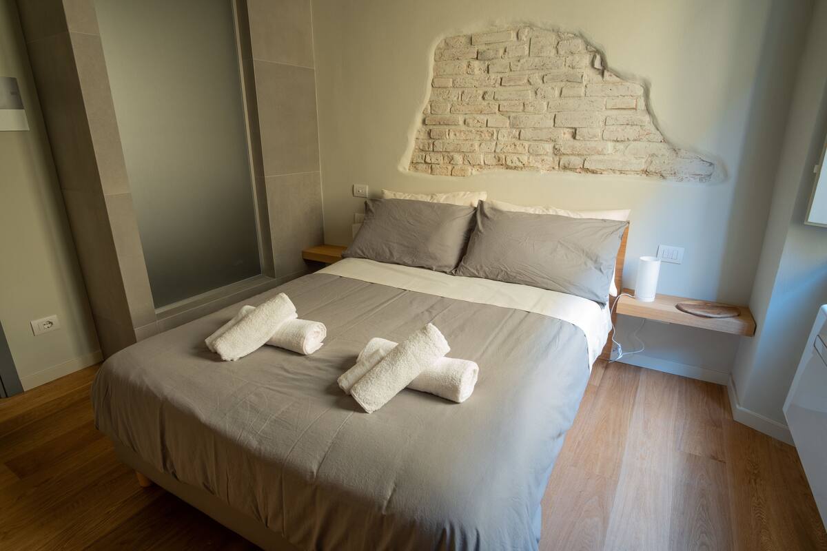 A neatly made bed with beige linens and two rolled towels, a brick-patterned wall, a bedside table with a lamp, and a wooden floor.
