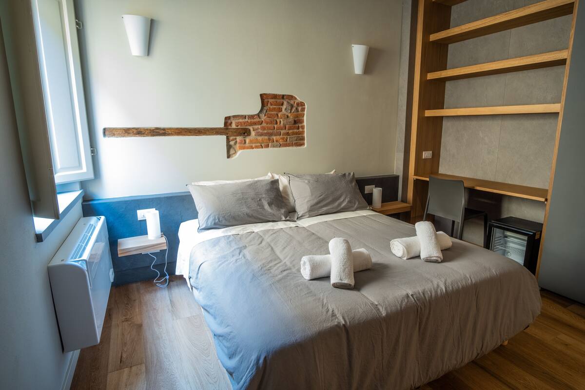 A neatly made bed with a grey duvet, two pillows, and two rolled towels. There is a brick wall decoration above the bed, a small bedside table with a lamp, and a heater on the wall.