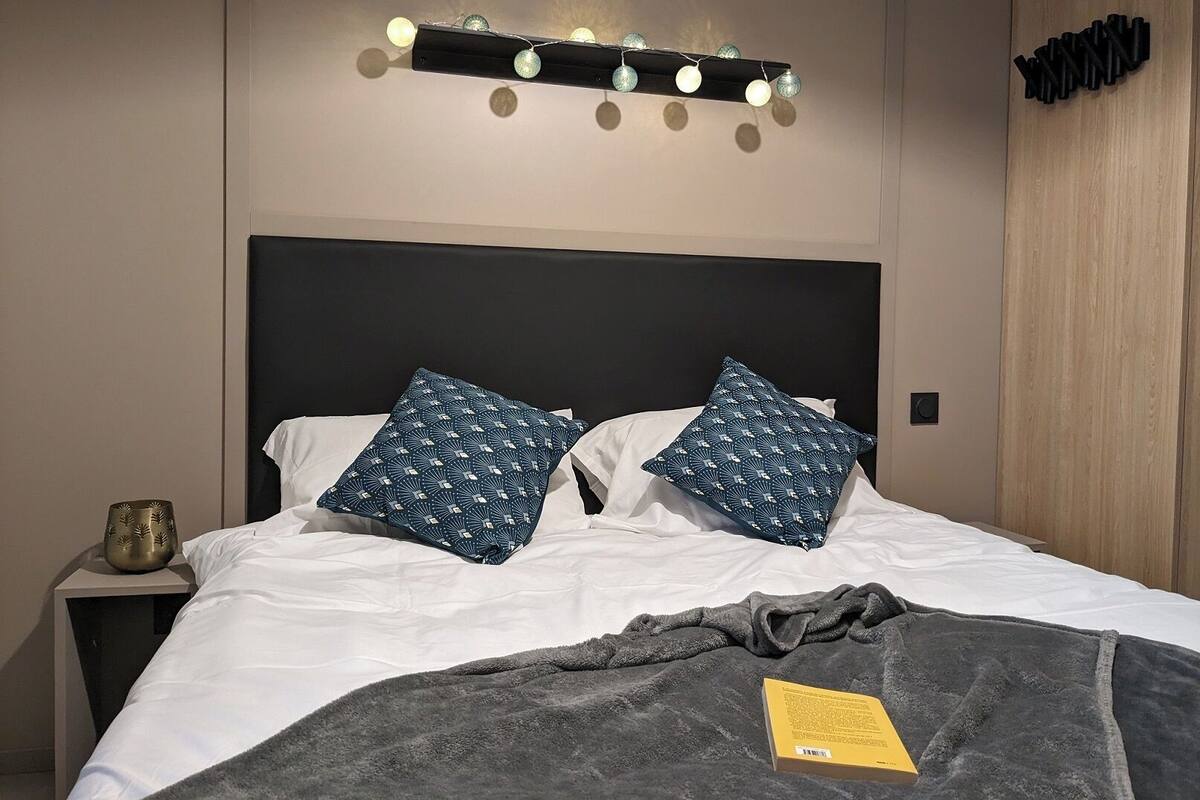 Een net opgemaakt bed met twee gekleurde kussens, een nachtkastje met een decoratief object en een wandlamp.