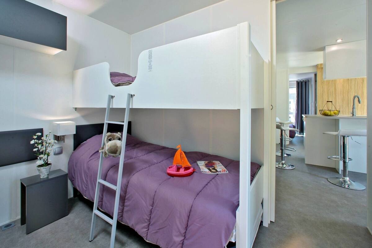Een moderne hotelkamer met een stapelbed, een klein bureau met lamp, een vaas met bloemen en uitzicht op de keuken.