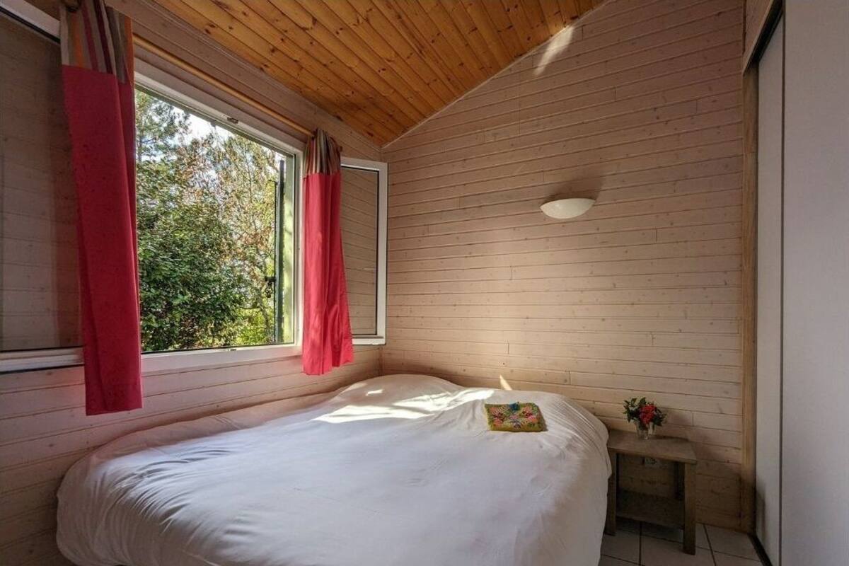Een slaapkamer met een houten plafond, een bed met witte beddengoed, een klein nachtkastje met een vaas, en een raam met rode gordijnen.
